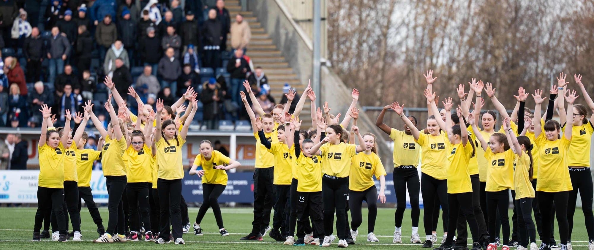 Stand-Out students in yellow t-shirts performing on the pitch at Falkirk Stadium with the crowd behind them