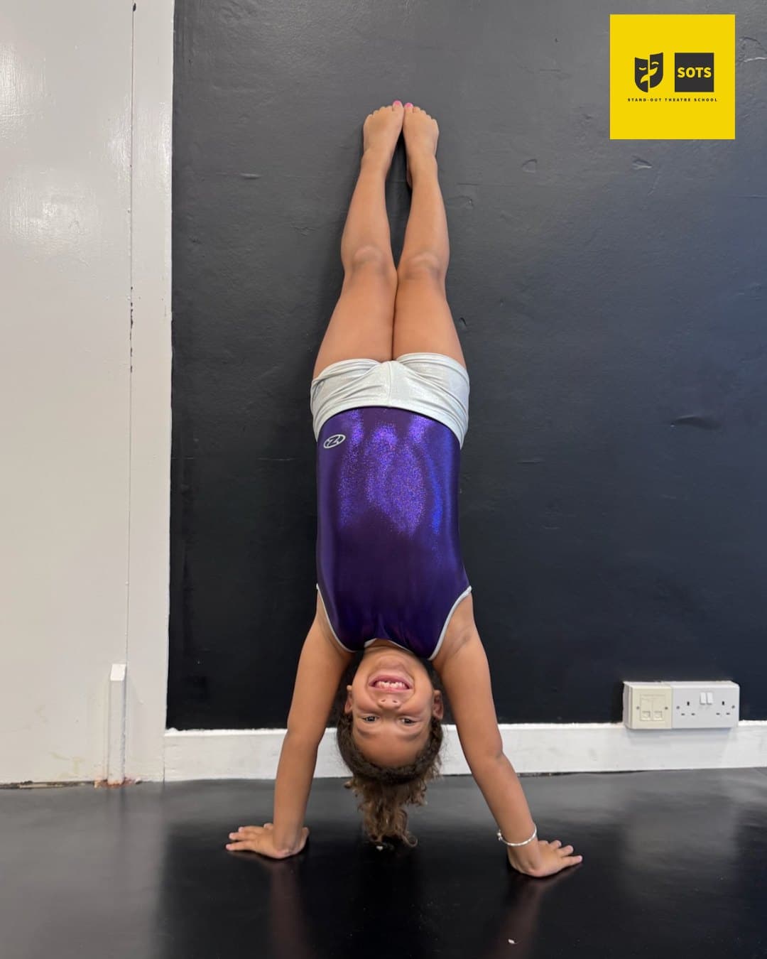 Mini Acro student in a purple leotard holding a handstand and smiling in the studio